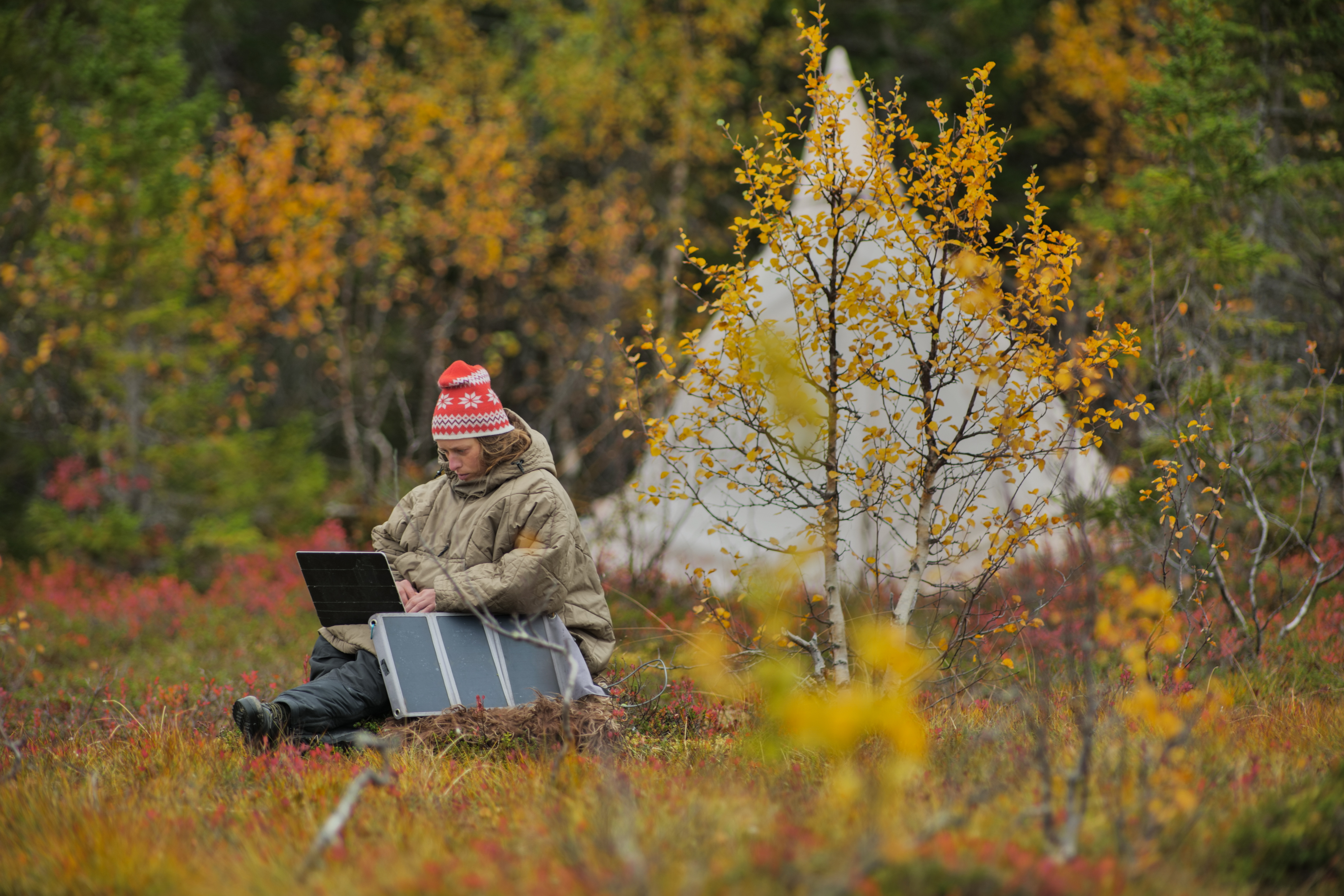caucasian female hiker sitting on a rock while working on a laptop being charged by solar panels nearby, portable solar green technology concept, camping in a beautiful autumn nature