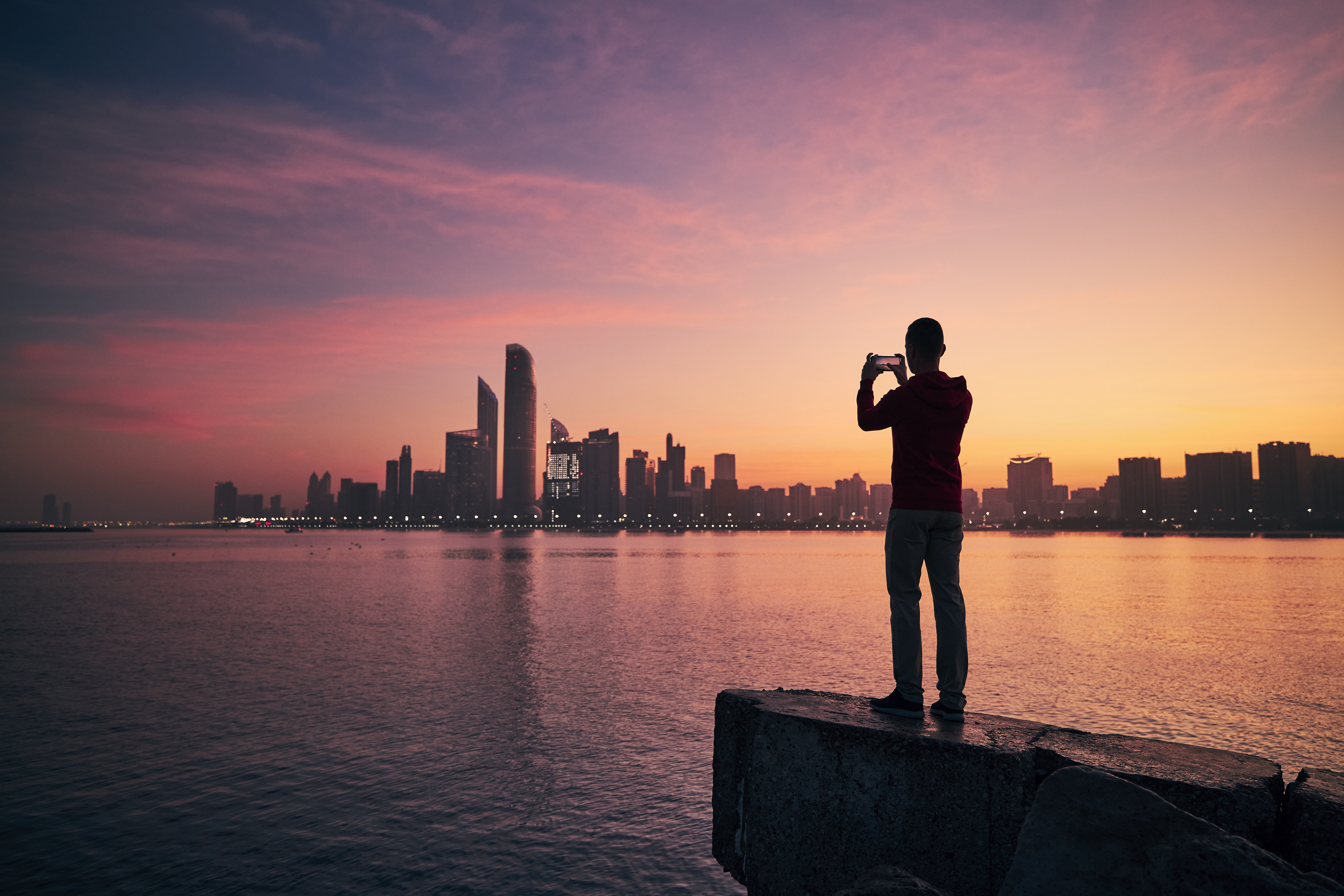 Young man with mobile phone photographing urban skyline at sunrise. Abu Dhabi, United Arab Emirates.